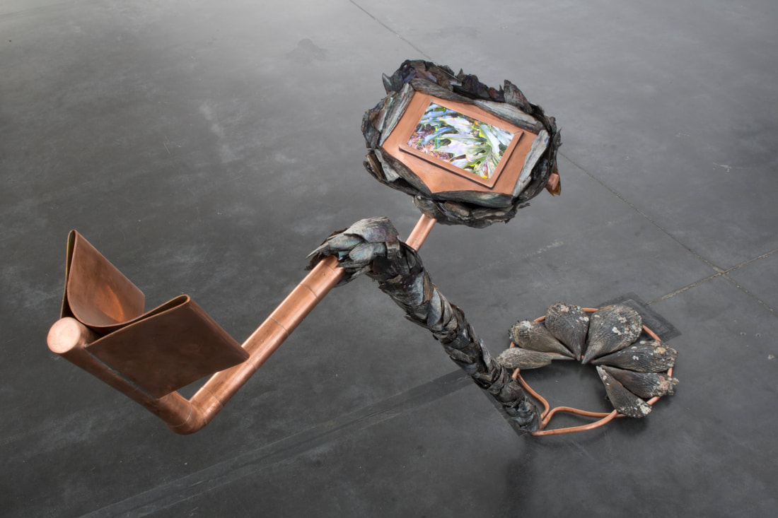  Indiscriminate Collector is a polished copper and shell sculpture in the shape of a metal detector. It has a screen portion that holds a monitor, playing a video of iridescent plants.